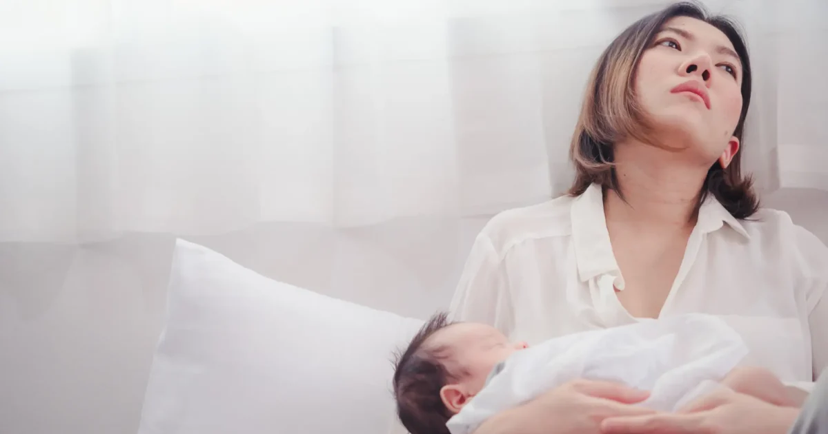 Valkopaitainen äiti istuu sängyllä ja pitelee vauvaa sylissään, katsoen väsyneesti ylöspäin kirkkaasti valaistussa huoneessa